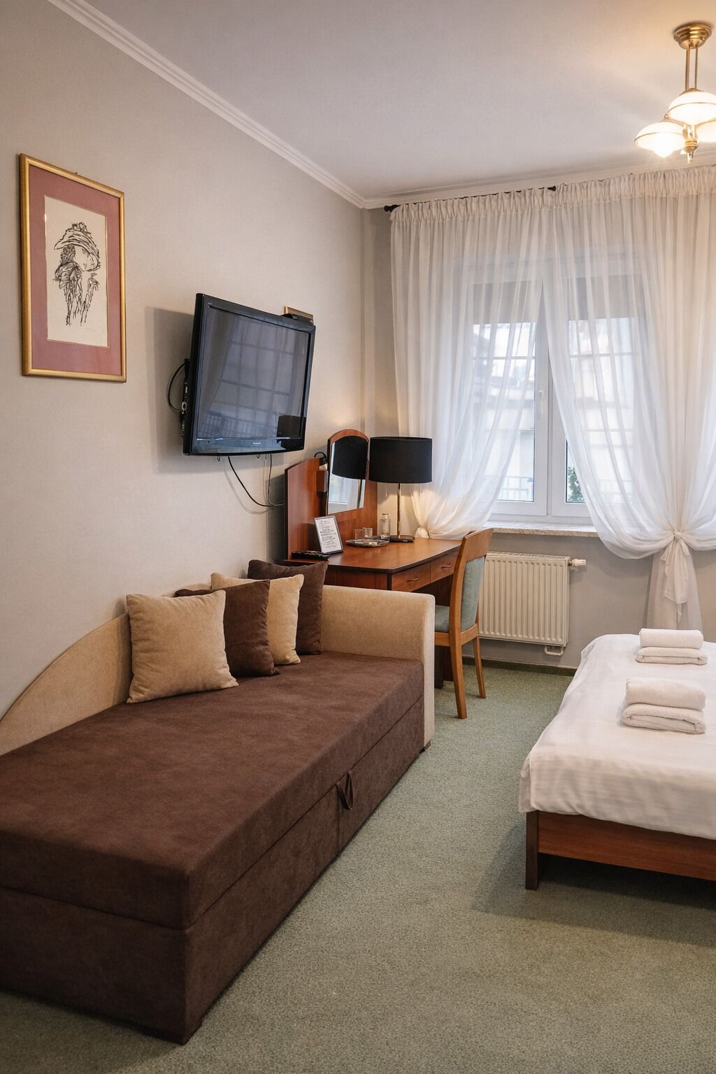 Una habitación de hotel con un sofá marrón, una cama con ropa blanca, un escritorio con computadora y lámpara, un televisor en la pared y cortinas traslúcidas.