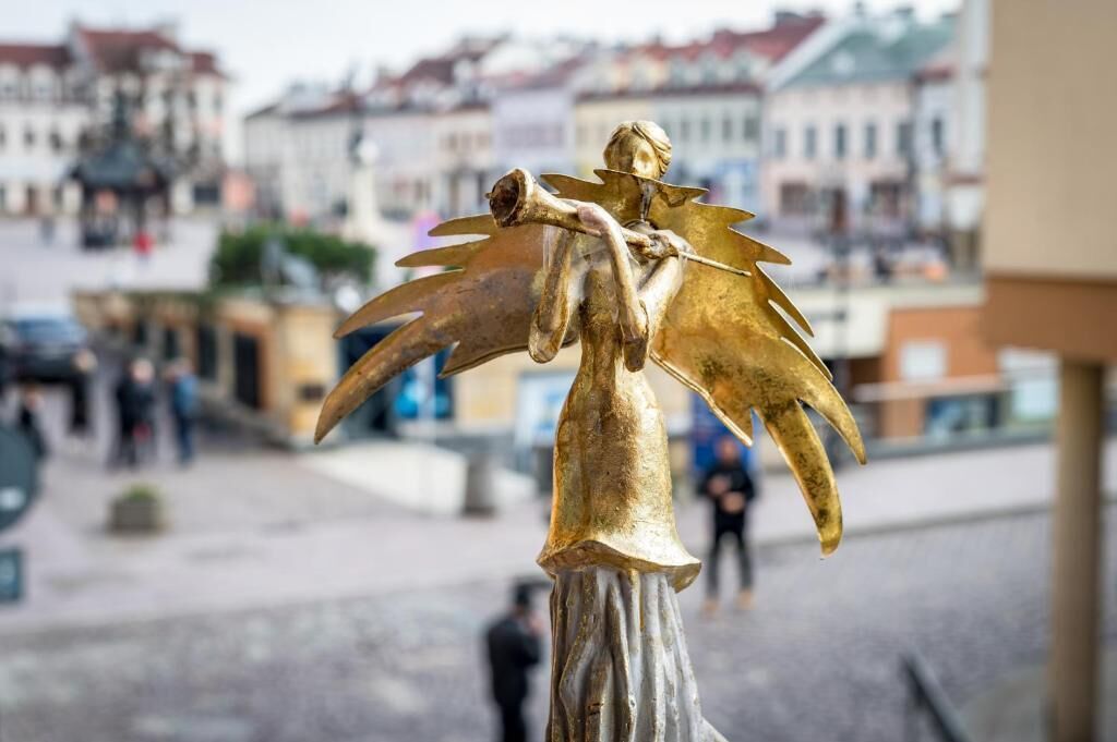 Une statue d’ange doré se dresse sur une place publique, avec des bâtiments en arrière-plan.
