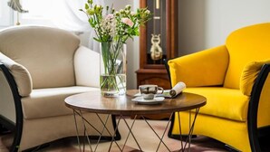 A room with a beige sofa, a yellow armchair, a round wooden table with a vase of flowers, a small cup, and a book. - Pod Ratuszem (Rzeszów)