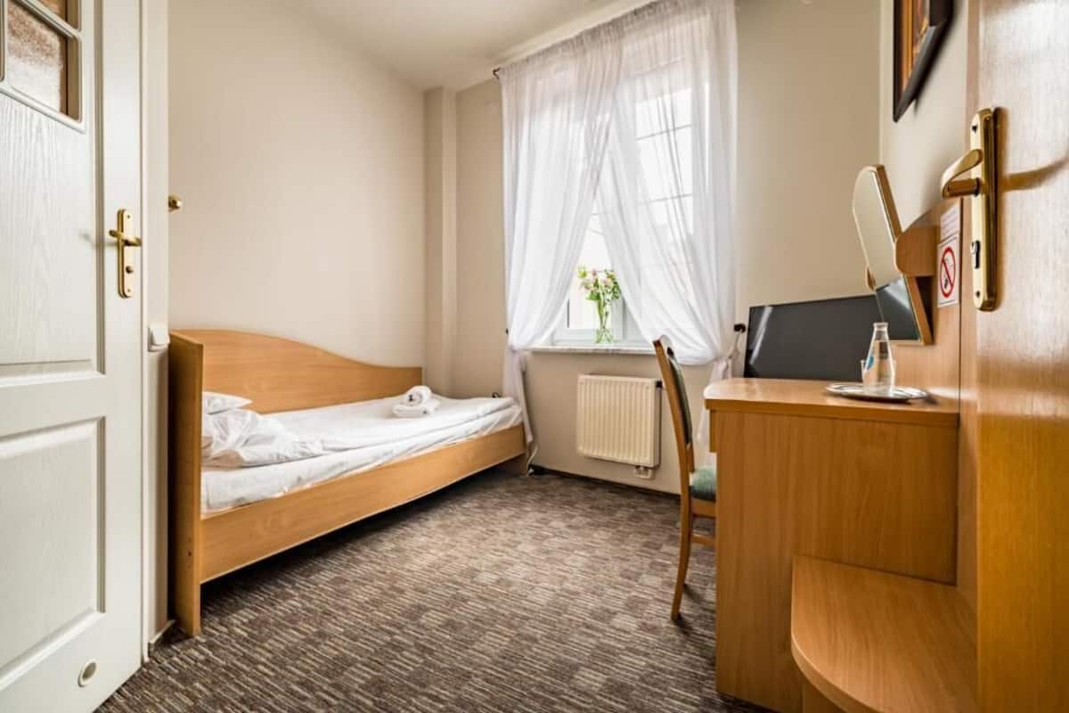 A small hotel room with a single bed, a desk with a computer, a chair, and a window with sheer curtains.