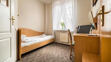 A small hotel room with a single bed, a desk with a computer, a chair, and a window with sheer curtains.