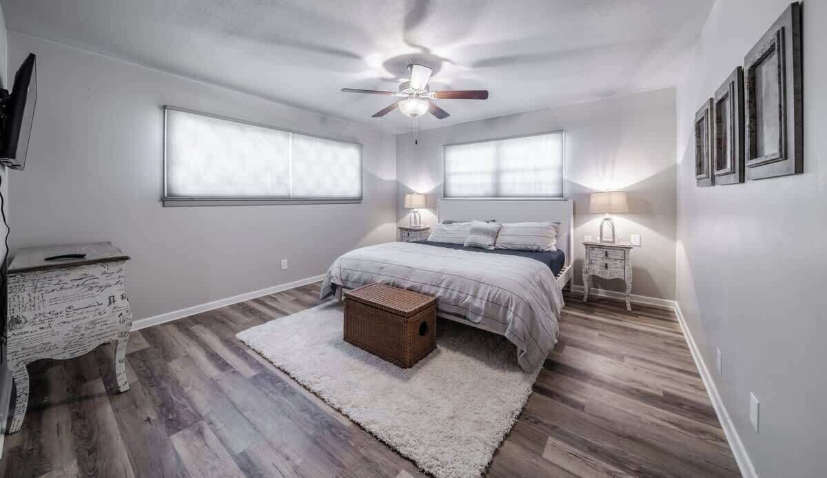 A bedroom with a bed, a ceiling fan, two bedside lamps, a small chest of drawers, and a large window.