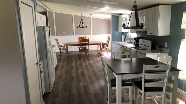 Una cocina con suelo de madera, armarios blancos, una mesa de comedor con sillas y una lámpara colgante.