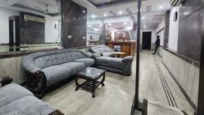 A hotel lobby with a curved gray sofa, a small wooden table, and a reception desk. - Hotel Mid Town (New Delhi)