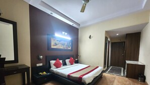 A hotel room with a bed, a desk, a chair, a nightstand, a wall-mounted lamp, a ceiling fan, and a door leading to another room. - Hotel Mid Town (New Delhi)