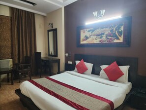Hotellihuone, jossa on suuri sänky, kaksi punaista tyynyä, puinen pääty, työpöytä tuolilla, suuri peili ja seinälle ripustettu taulu.