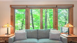 A room with a grey sofa, two nightstands with lamps, and large windows with a forest view.
