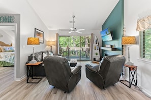 A modern living room with a ceiling fan, two leather chairs, a sofa, a dining table with chairs, and a balcony with a view of trees.