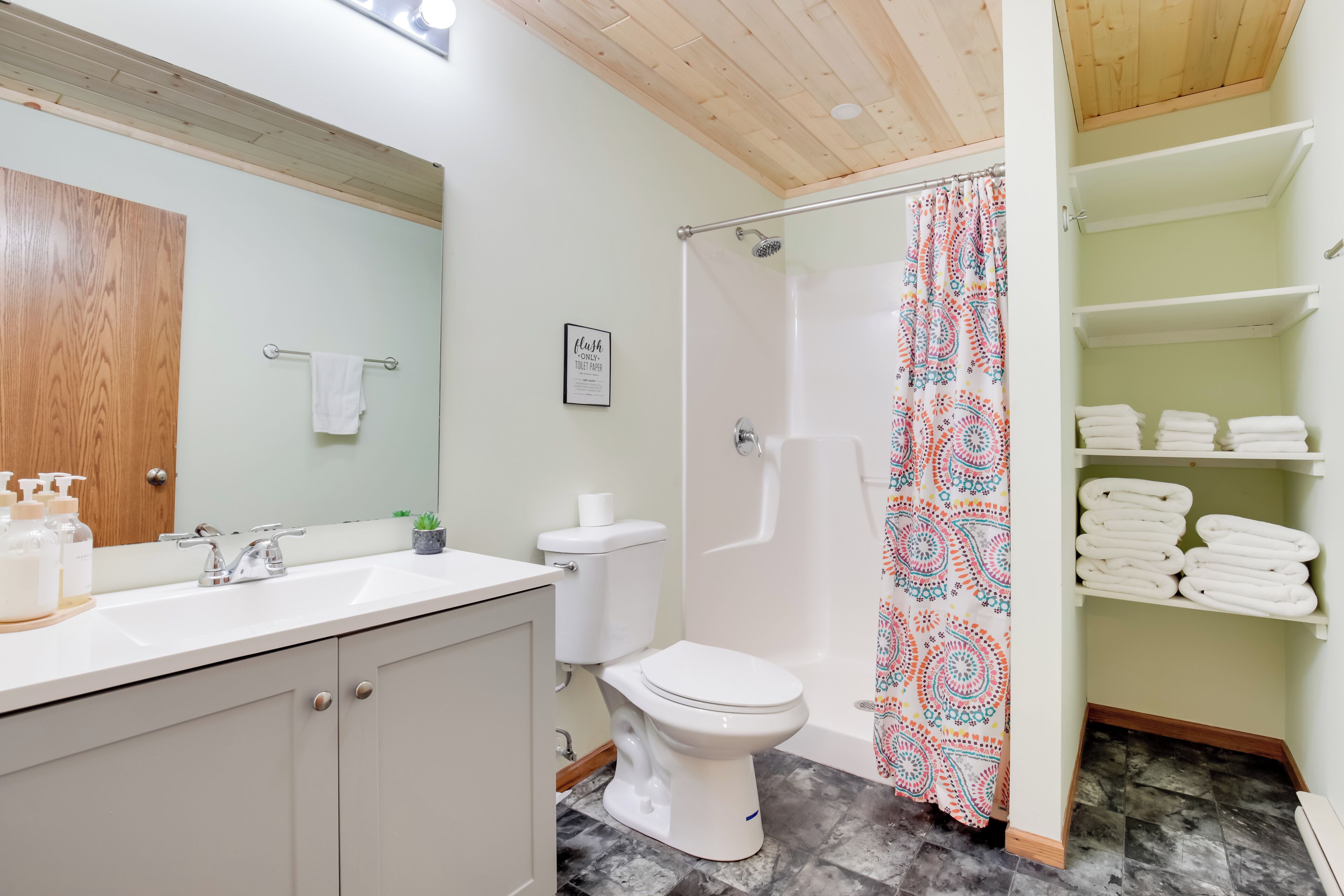 Een badkamer met een houten plafond, een witte wastafel, een toilet en een douche met een decoratieve gordijn. Handdoeken liggen op planken en er hangt een bordje aan de muur.