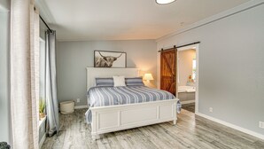 Un dormitorio con cama, mesita de noche, lámpara, un cuadro enmarcado en la pared y una puerta de madera que da a otra habitación.