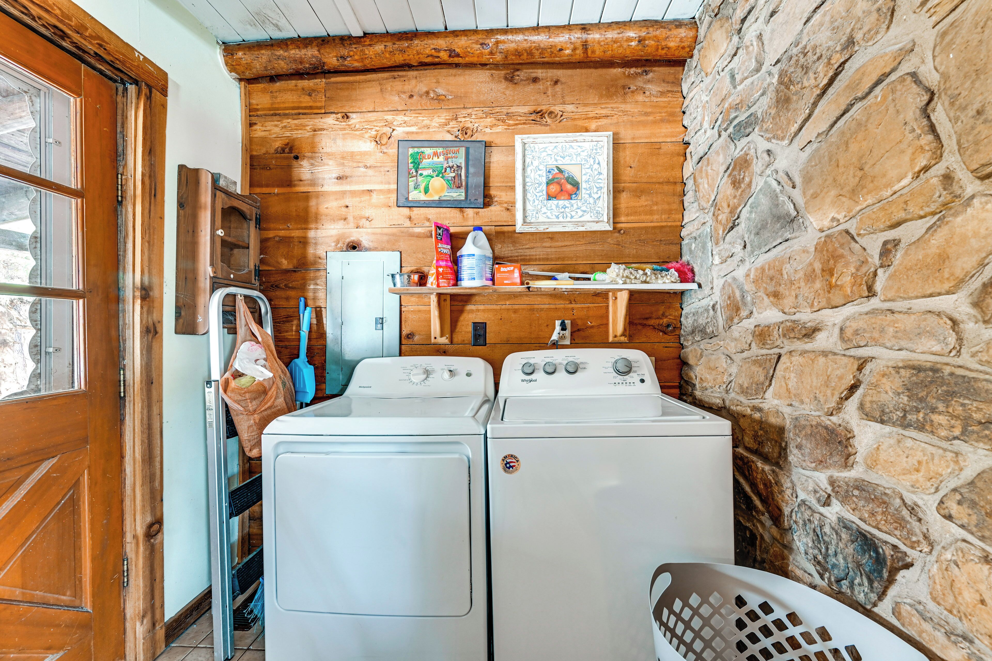 A laundry room with two white front-loading washers and dryers, wooden walls, a stone accent wall, and a shelf with various items.