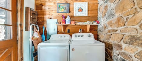 A laundry room with two white front-loading washers and dryers, wooden walls, a stone accent wall, and a shelf with various items.
