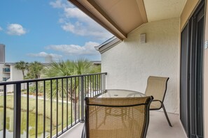 A balcony with a glass table and wicker chairs, overlooking a landscaped area with palm trees and a building in the background.