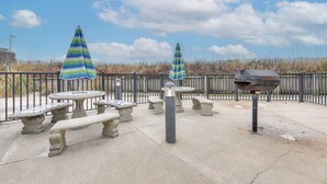 Un patio al aire libre con mesas y bancos de piedra, dos sombrillas a rayas y una parrilla.
