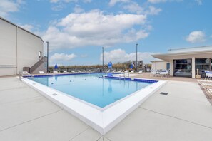 A rectangular swimming pool with a white border and blue tiles, surrounded by white lounge chairs and a covered seating area.