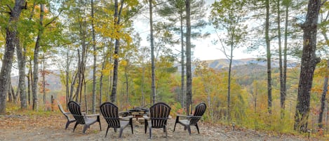 A rustic outdoor seating area with four Adirondack chairs around a wooden table, set in a forested area with autumn foliage.