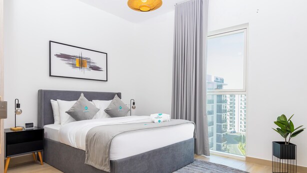 Une chambre moderne avec un lit gris, du linge de lit blanc, une œuvre d’art encadrée, une table de chevet avec une lampe et une vue sur le paysage urbain à travers une grande fenêtre.