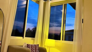 Ein gemütliches Zimmer mit einer Couch, einer Lampe und großen Fenstern mit Vorhängen.