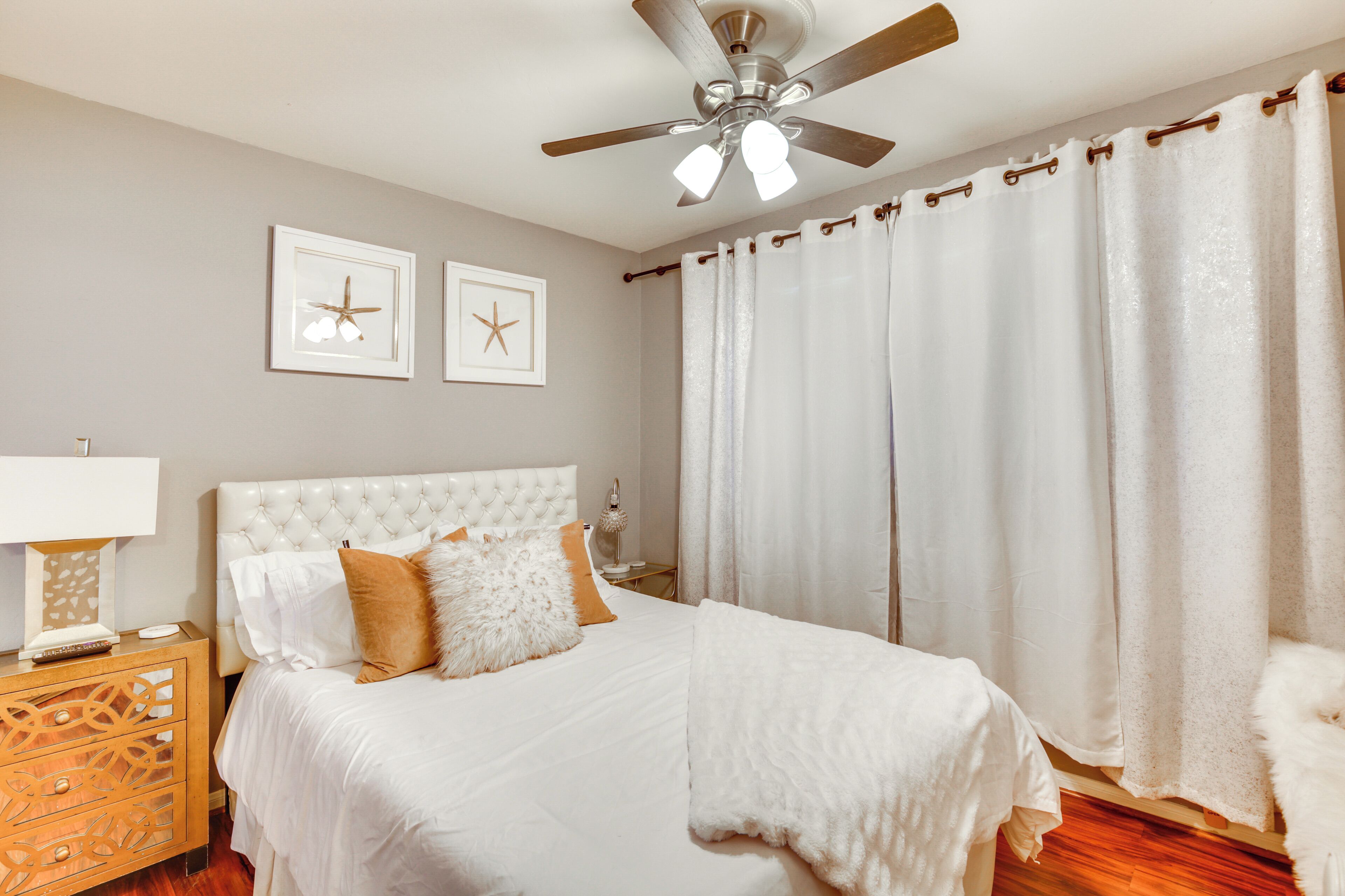 A bedroom with a tufted headboard, a bed with white bedding, a wooden nightstand with a lamp, and a ceiling fan.