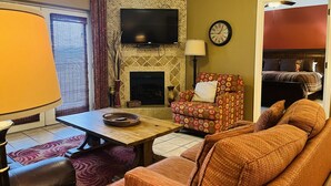 Een woonkamer met een open haard met bakstenen patroon, een aan de muur bevestigde televisie, een bank, een fauteuil met een patroon en een eettafel met een lamp.