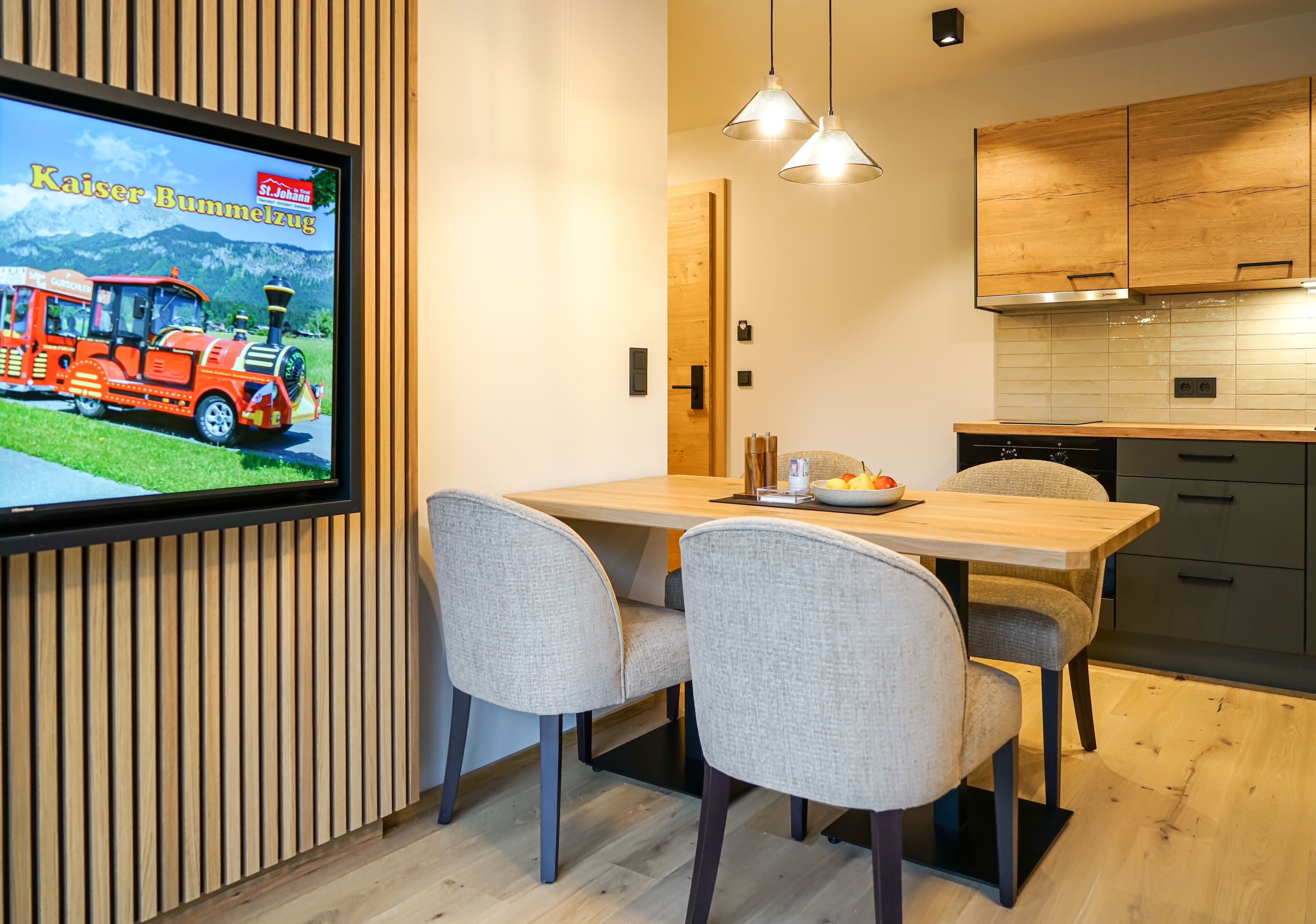 A modern kitchen with wooden cabinets, a dining table with chairs, and a television displaying a train advertisement.