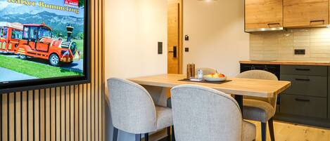 A modern kitchen with wooden cabinets, a dining table with chairs, and a television displaying a train advertisement.