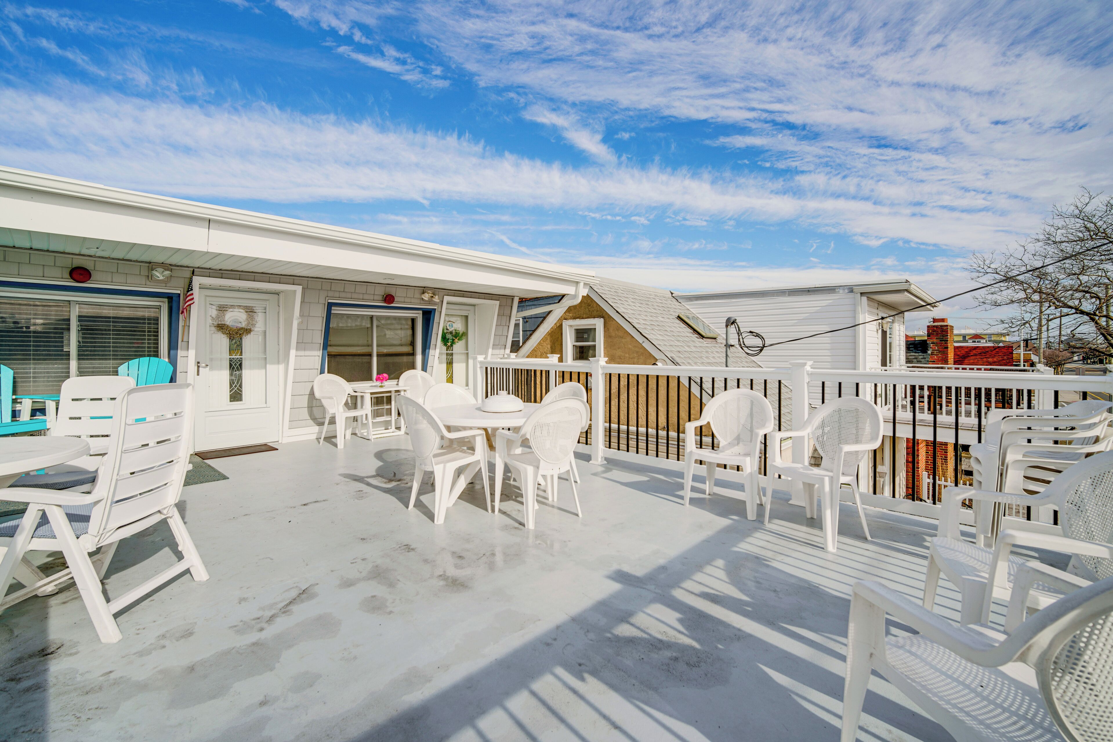 A covered outdoor patio with white plastic chairs and tables, a white fence, and a view of the street.
