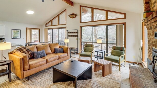 A spacious living room with a leather sofa, a coffee table, and a fireplace. There are multiple windows providing natural light and a view of the outdoors. - Premier Trout Fishing: Scenic White River Cabin (Lakeview)