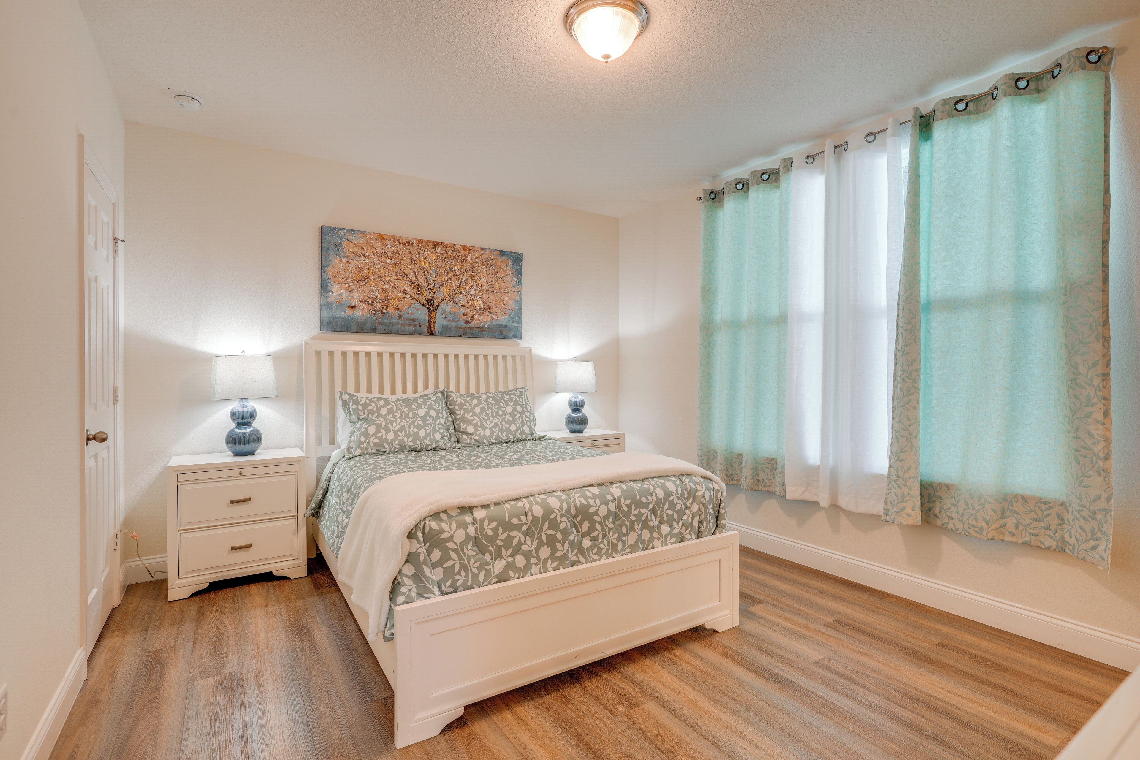 A bedroom with a bed, nightstands, a lamp, a painting, and curtains.
