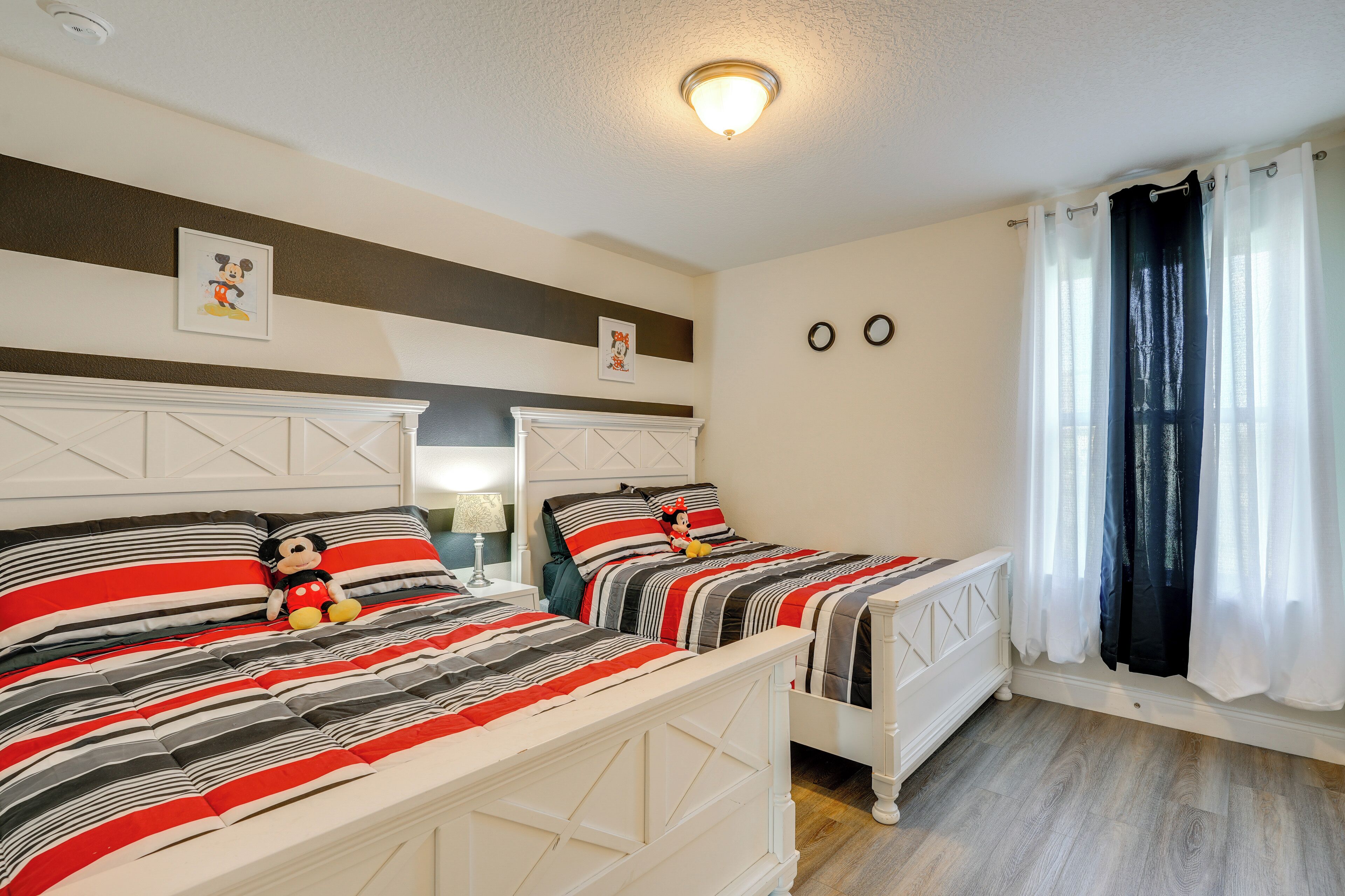 Een slaapkamer met twee bedden, streepjesbeddengoed, een Mickey Mouse knuffel, en een nachtkastje met een lamp.