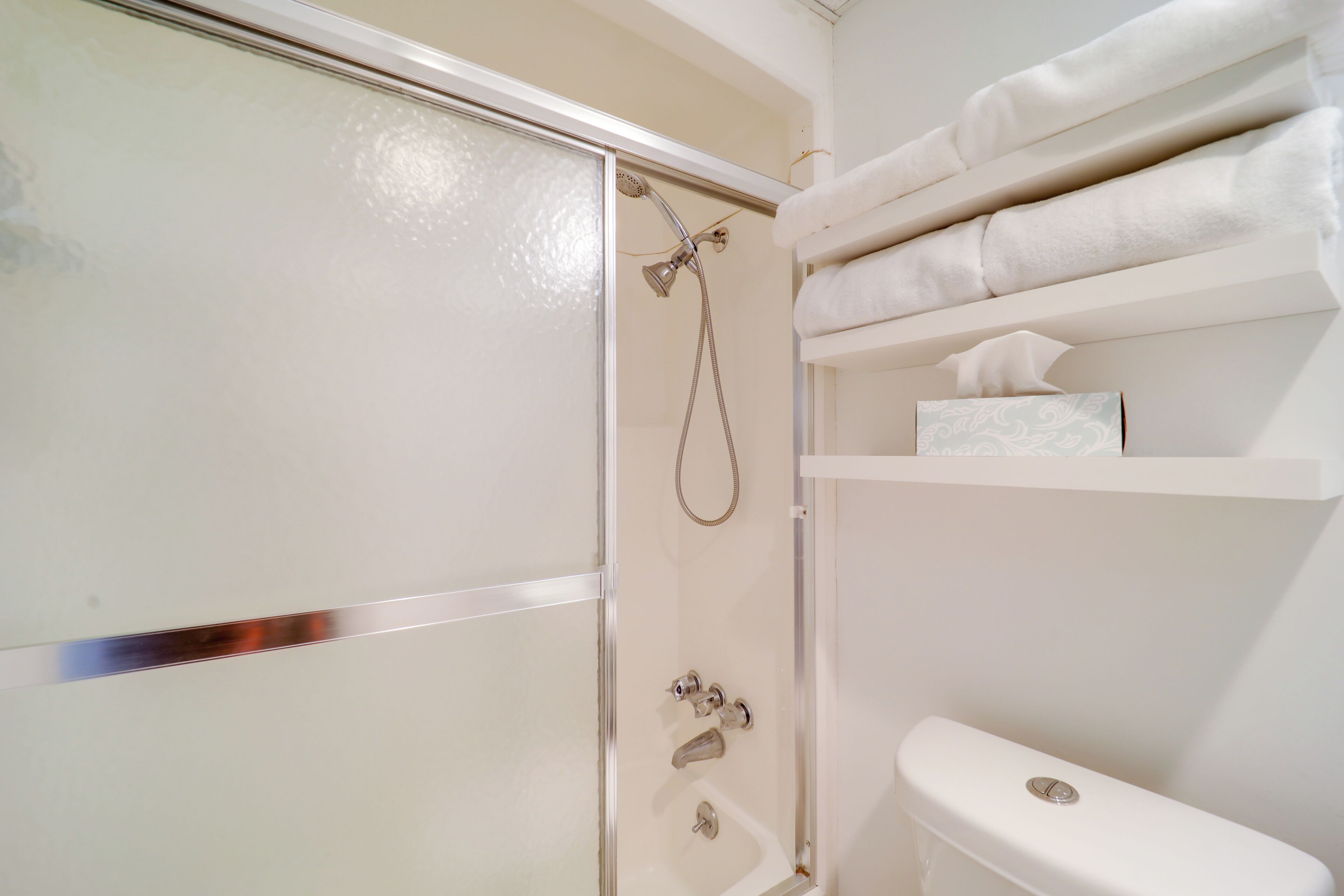 A bathroom with a glass shower enclosure, a toilet, and shelves with towels and a tissue box.