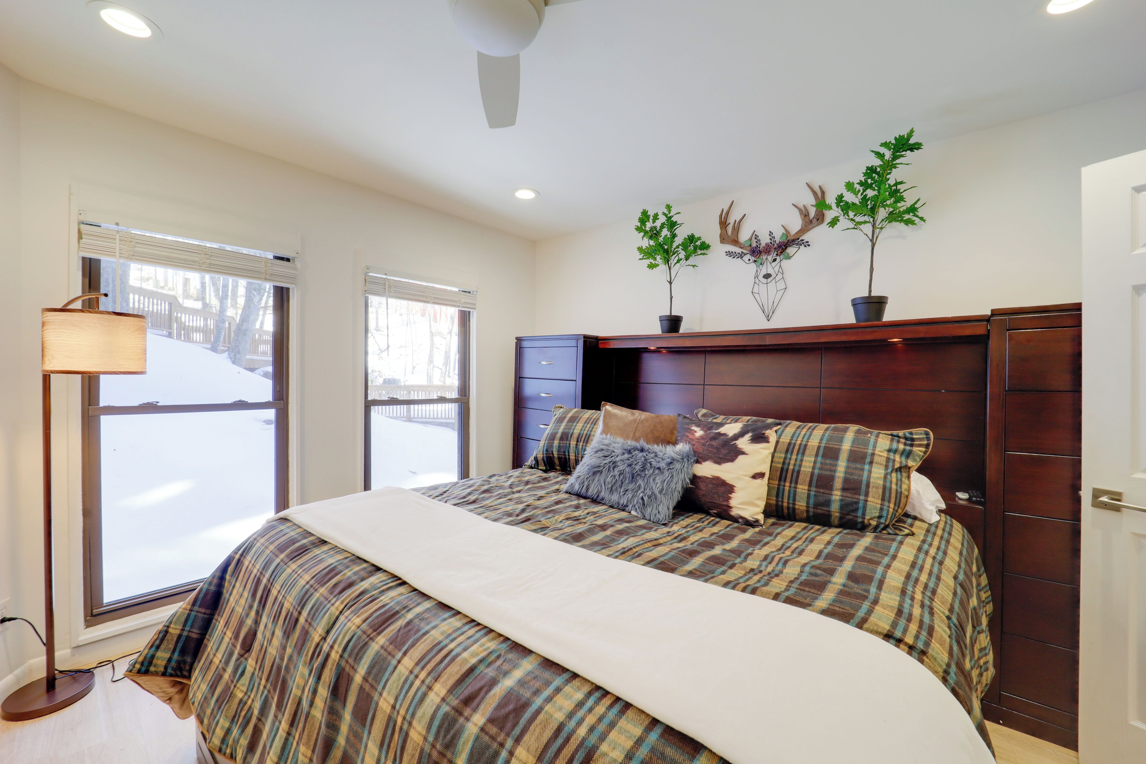 A bedroom with a large bed, a wooden headboard, two windows with blinds, a floor lamp, and wall decorations including a plant and antlers.