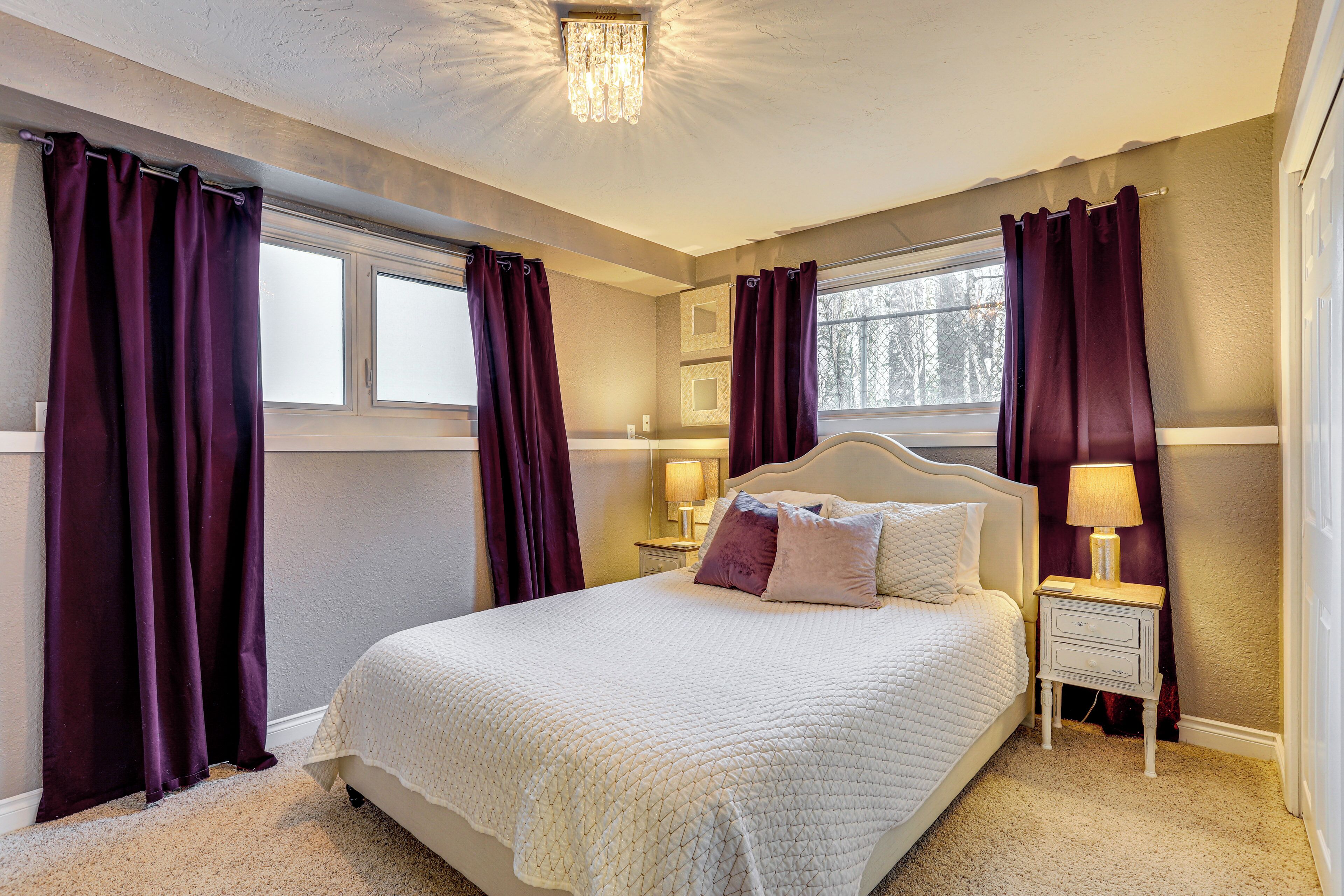 A bedroom with a large bed, two bedside tables, a window with curtains, and a chandelier.