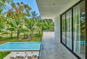 A modern poolside area with a clear view of a swimming pool, lounge chairs, and a glass balustrade overlooking a tropical garden with palm trees.