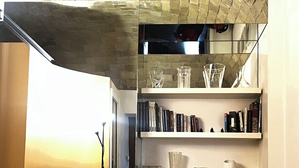 A bedroom with a brick wall, a bed with white linens, a TV on a side table, and shelves with books and glassware.