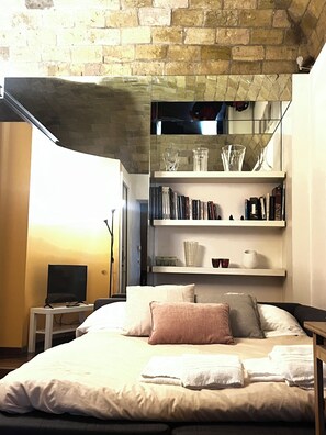 A bedroom with a brick wall, a bed with white linens, a TV on a side table, and shelves with books and glassware. - The Ancient Wall | Borgo Pio | San Pietro (Roma)