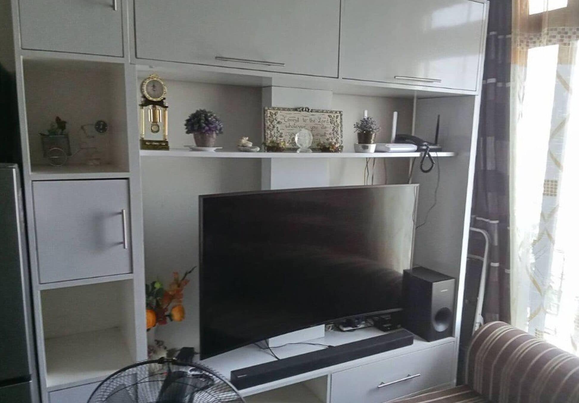 A living room with a flat-screen TV, a speaker, and a fan. There are shelves with decorative items and a clock.