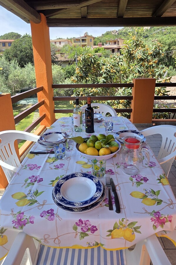 An outdoor dining area with a table set for two, featuring a floral tablecloth, a bowl of lemons, a pitcher, and wine glasses.