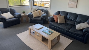 A modern living room with two black leather sofas, a wooden coffee table, and a view of the outdoors through large windows. - Conveniently located in the centre of town and close to the beaches (Bridport)