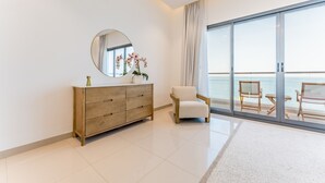 Ein modernes Zimmer mit einem Holzkasten, einem runden Spiegel, einem weißen Stuhl und einer großen Schiebeglastür, die auf einen Balkon mit Meerblick führt.