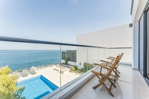 A modern balcony with a pool, wooden chairs, and a view of the sea.