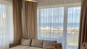 Un salon moderne avec un canapé d’angle, une table à manger et des chaises. La pièce est dotée de grandes fenêtres avec des rideaux légers et offre une vue sur la plage.