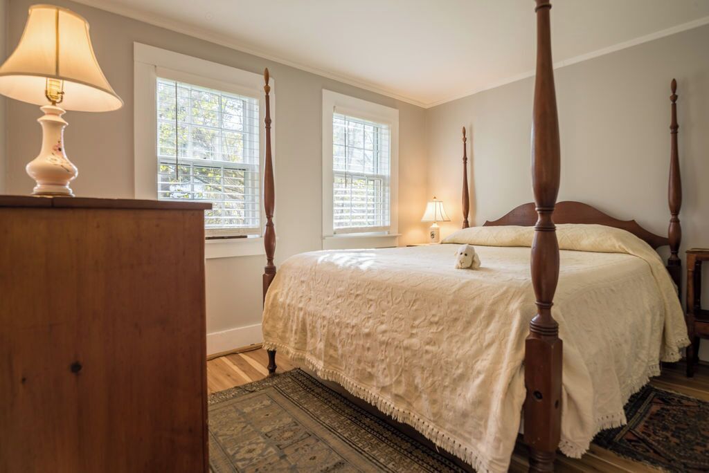 A bedroom with a four-poster bed, two windows, a nightstand, a lamp, and a rug.