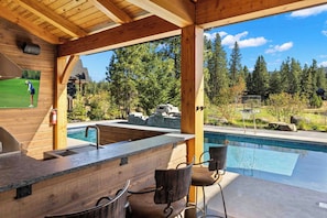 Un espace bar extérieur en bois, doté d’une piscine, de tabourets de bar et offrant une vue sur un terrain de golf.