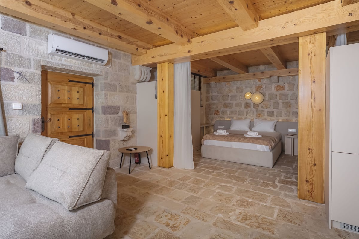 A cozy room with a stone wall, wooden beams, a bed, a small table, and a sofa.