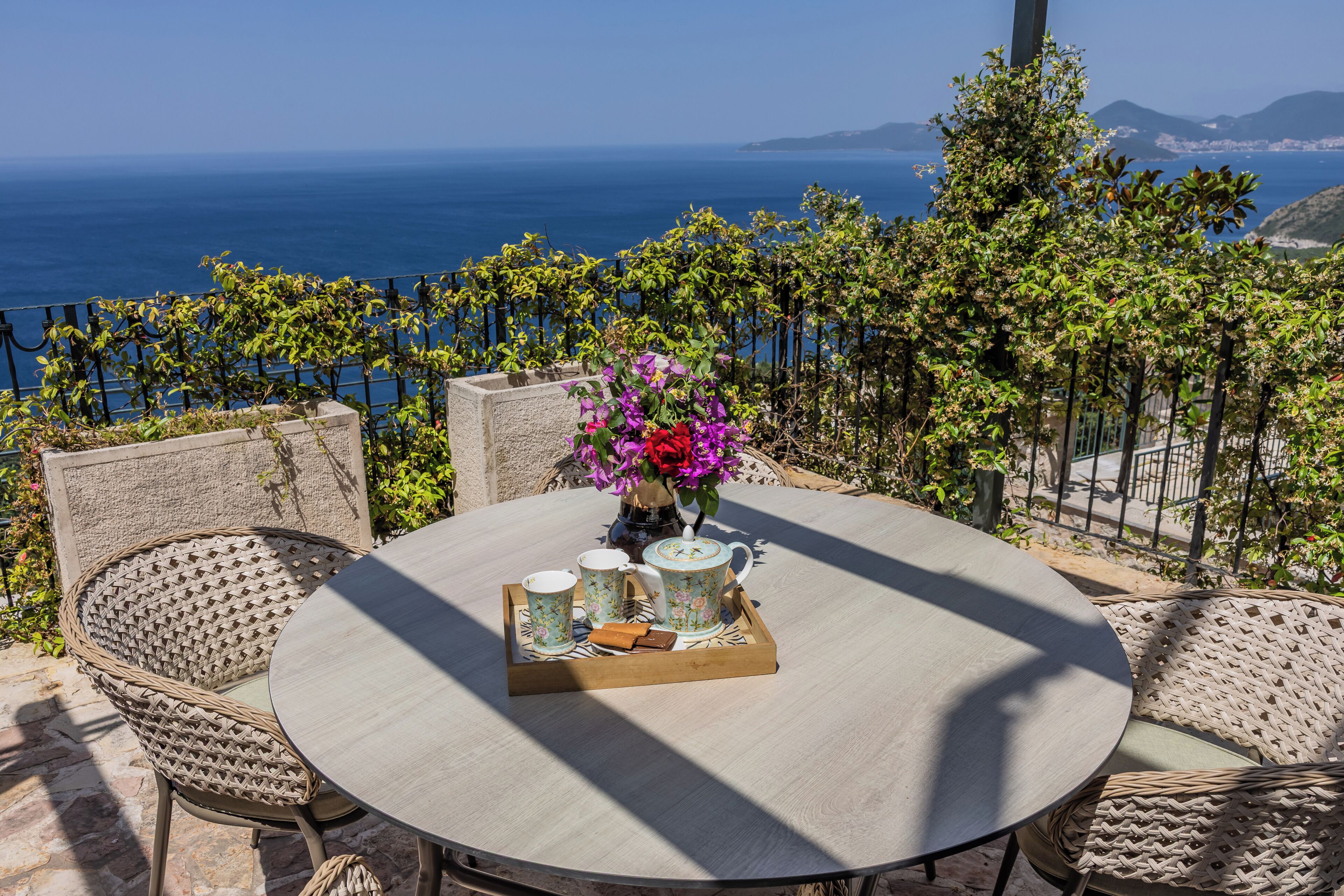 A round table with wicker chairs, a tray with teacups and a vase of flowers, and a view of the sea.