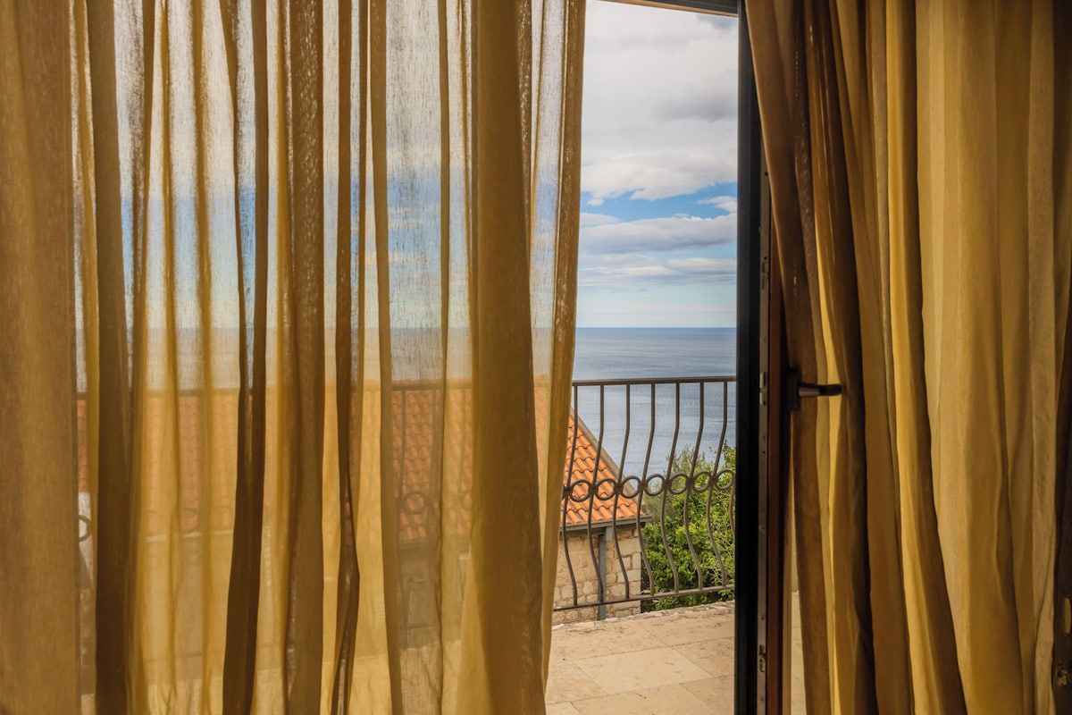 A view through a window with curtains, revealing a balcony with a railing and a building with a red roof in the background.
