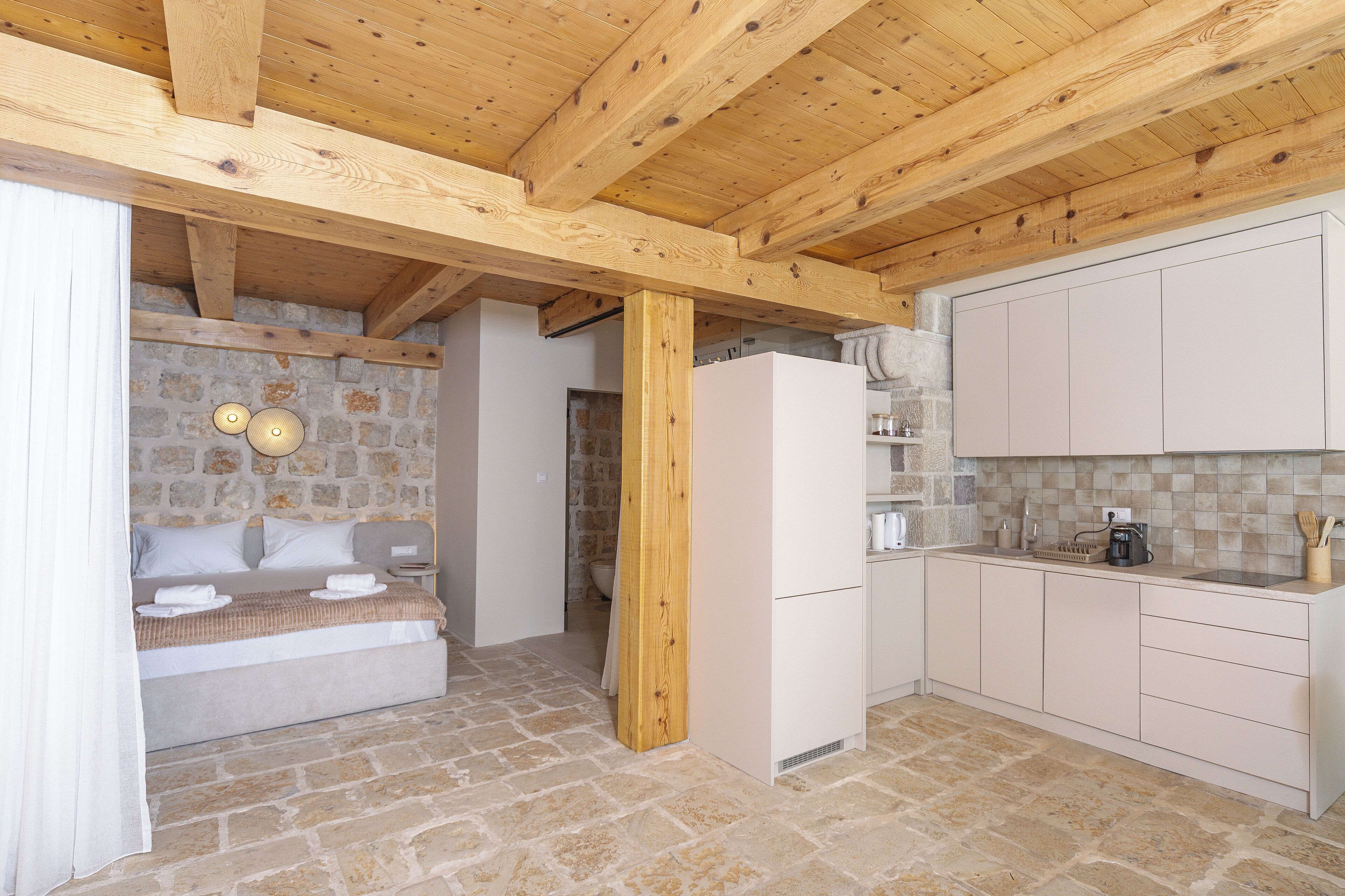 A room with a stone wall, wooden ceiling beams, a bed, a white refrigerator, and a kitchen area with cabinets and a countertop.