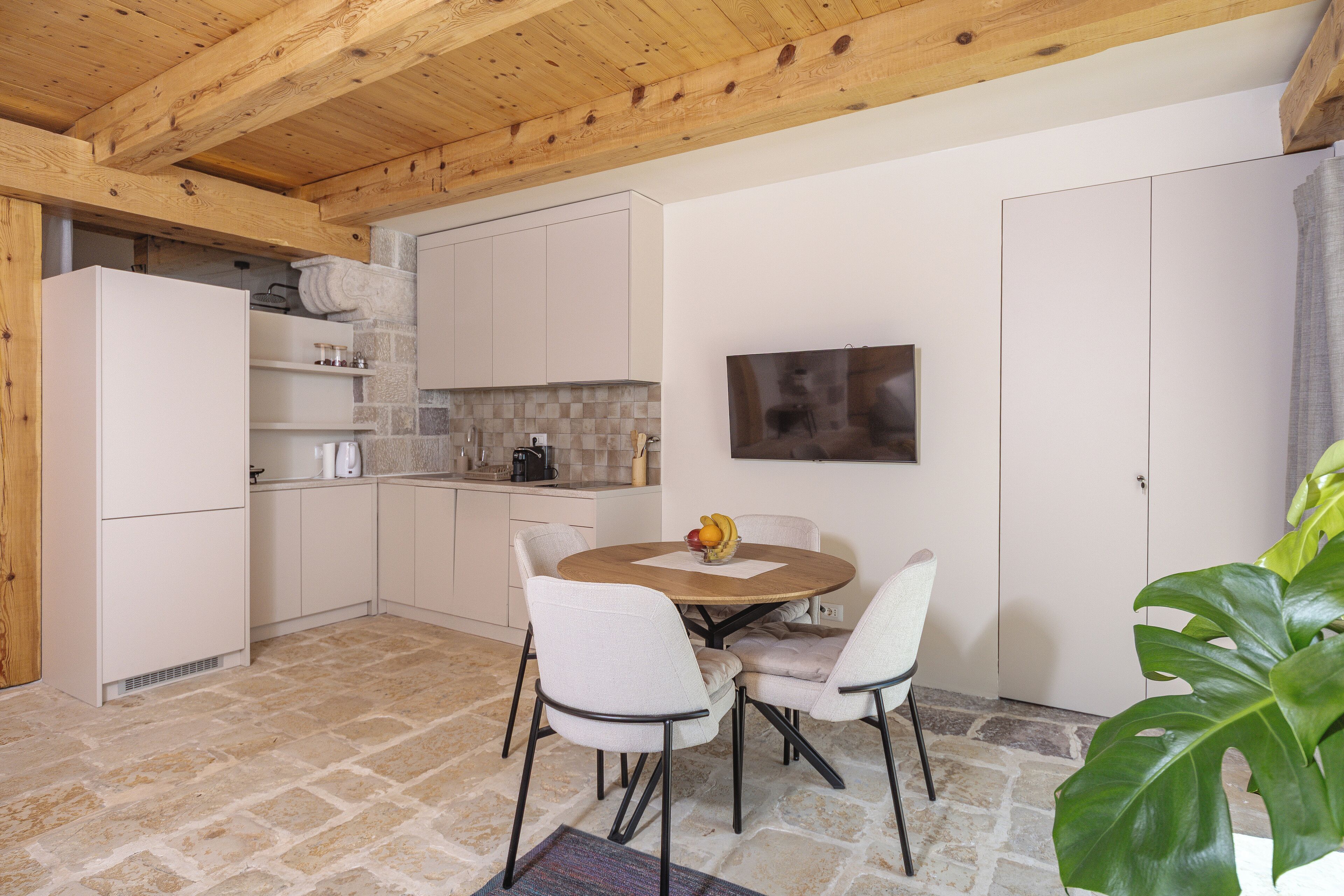 A modern kitchen with white cabinets, a stone backsplash, and a wooden ceiling. There is a dining table with chairs and a fruit bowl on it.