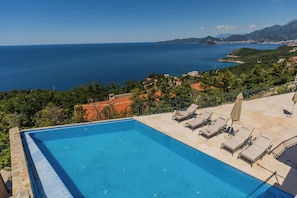 Ein rechteckiger Pool mit Liegestühlen und Sonnenschirmen, umgeben von einer Steinmauer und Blick auf das Meer und die Hügel.